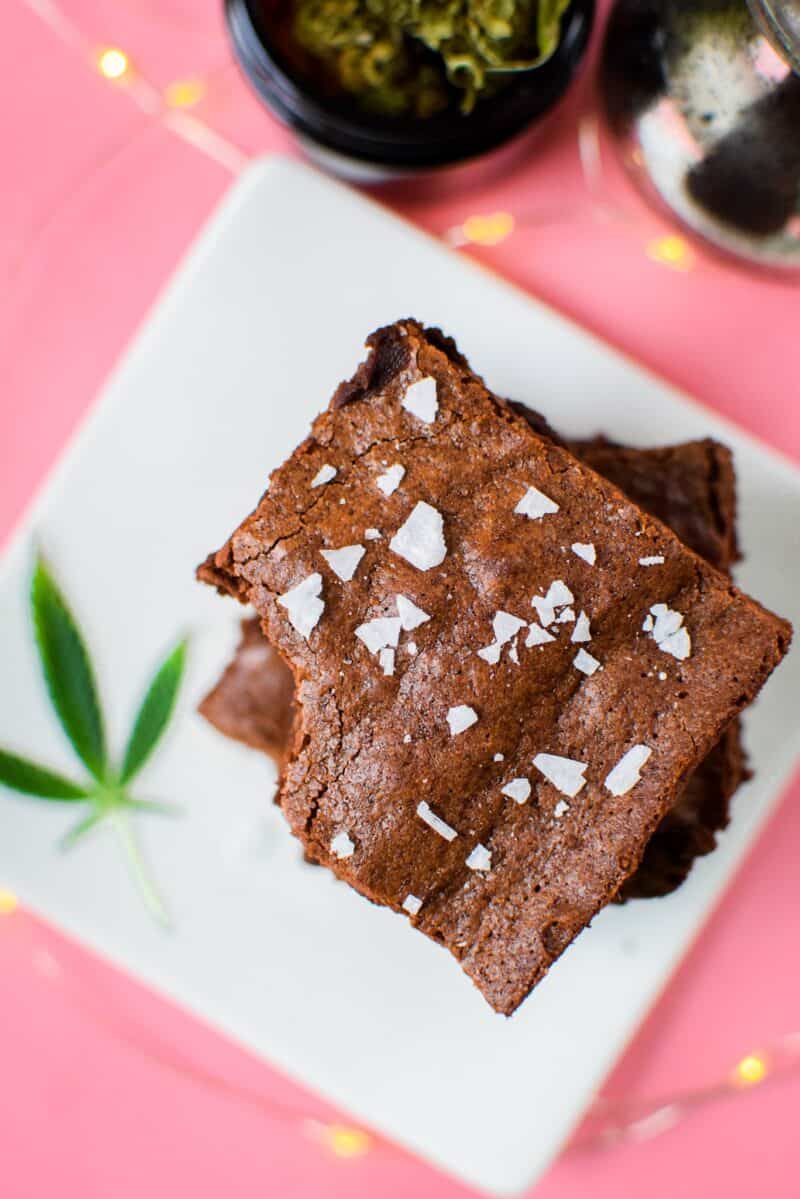 A white plate on a pink background with three cannabis brownies stacked on top, garnished with a cannabis leaf