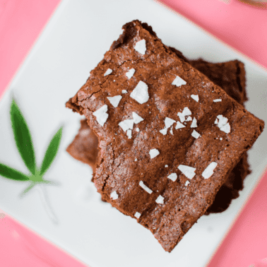 A white plate on a pink background with three cannabis brownies stacked on top, garnished with a cannabis leaf