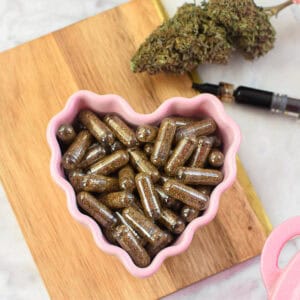 Cannabis capsules in a pink heart-shaped dish with cannabis buds and dropper.