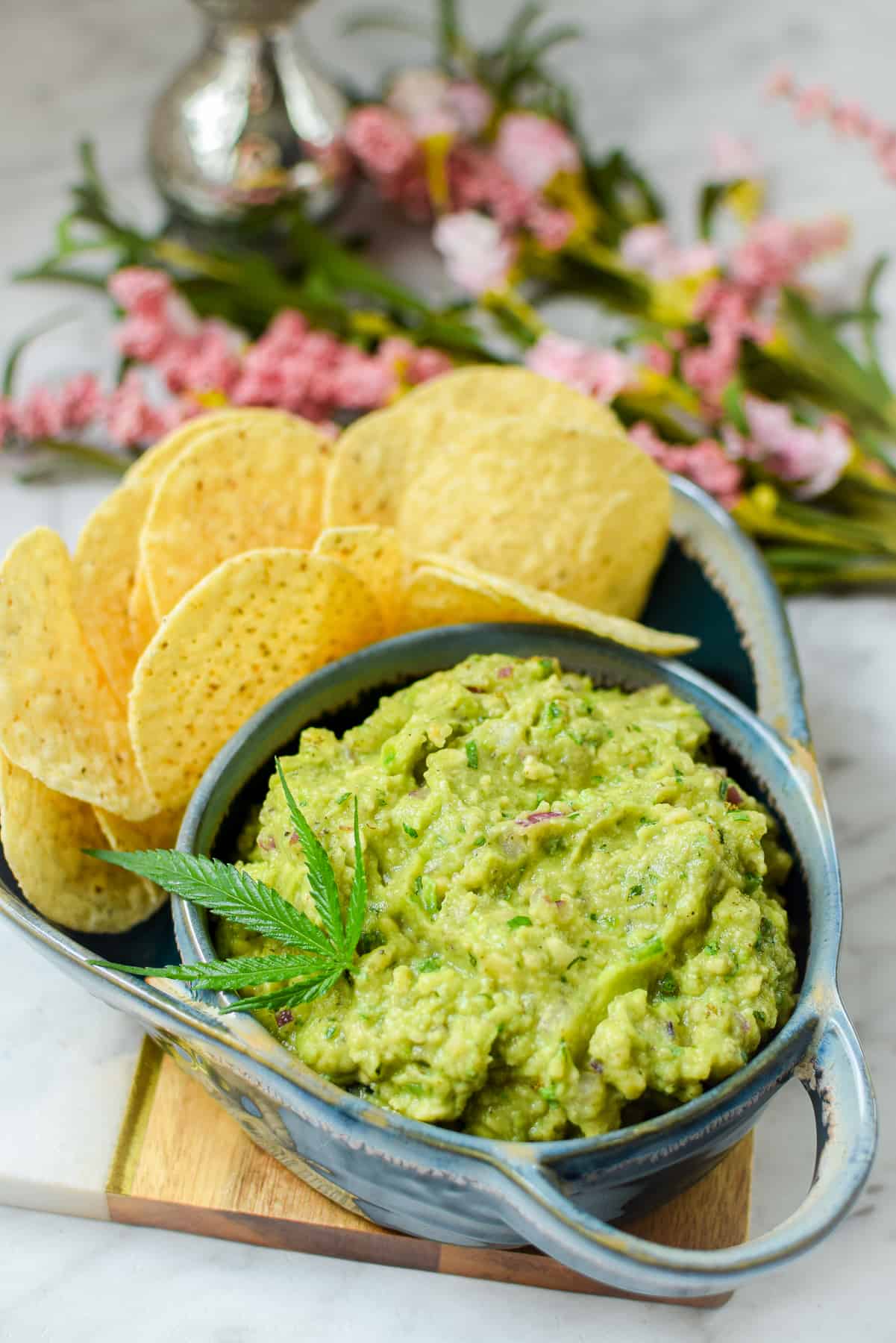 A blue bowl filled with guacamole garnished with a cannabis leaf paired with round tortilla chips