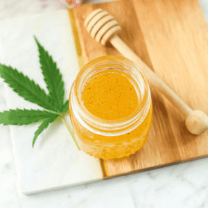 A finished image of cannabis-infused honey with pink flowers and a fresh cannabis leaf in the back, with a honey dobber about to be added to the jar.