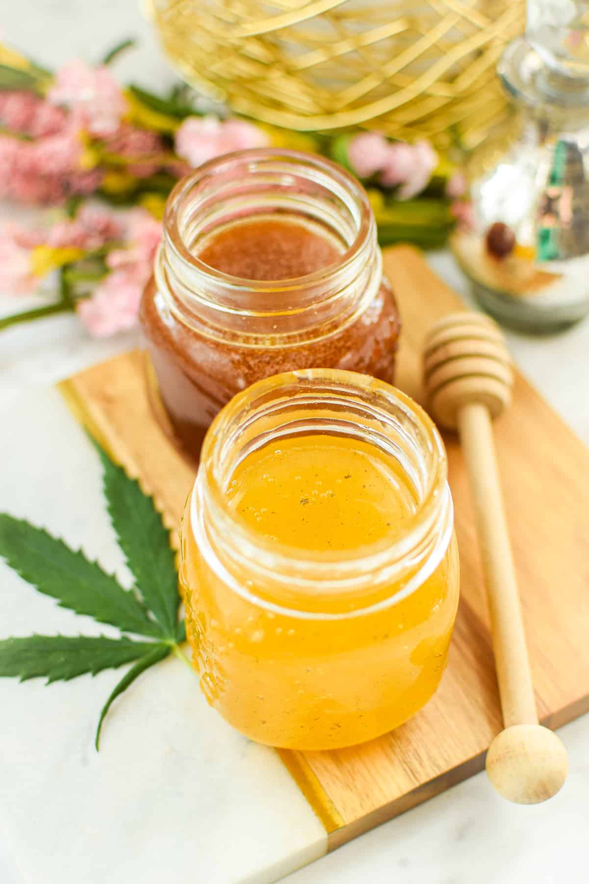 A finished image of cannabis-infused honey with pink flowers and a fresh cannabis leaf in the back, with a honey dobber on the side