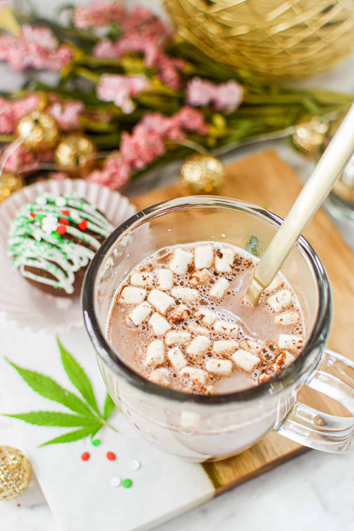 A white countertop with a cutting board topped with a clear mug full of milk and a hot cocoa bomb that has released cocoa mix and marshmallows