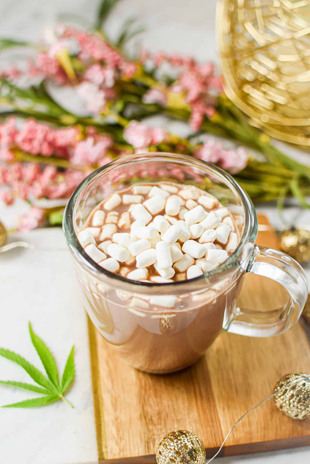 A white countertop with a cutting board topped with a clear mug full of cannabis hot cocoa with marshmallows