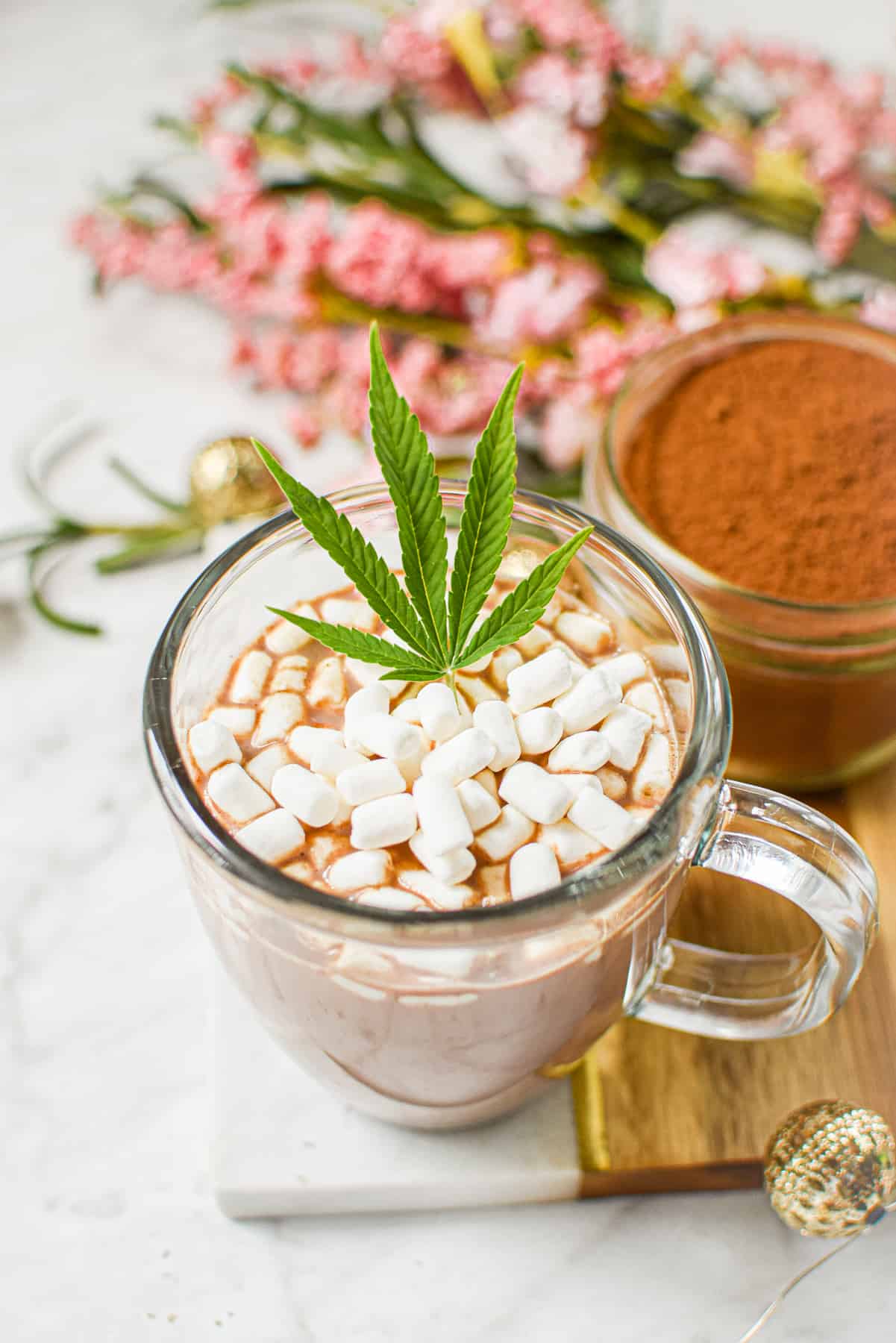 A white countertop with a cutting board topped with a clear mug full of cannabis hot cocoa with marshmallows, garnished with a cannabis leaf