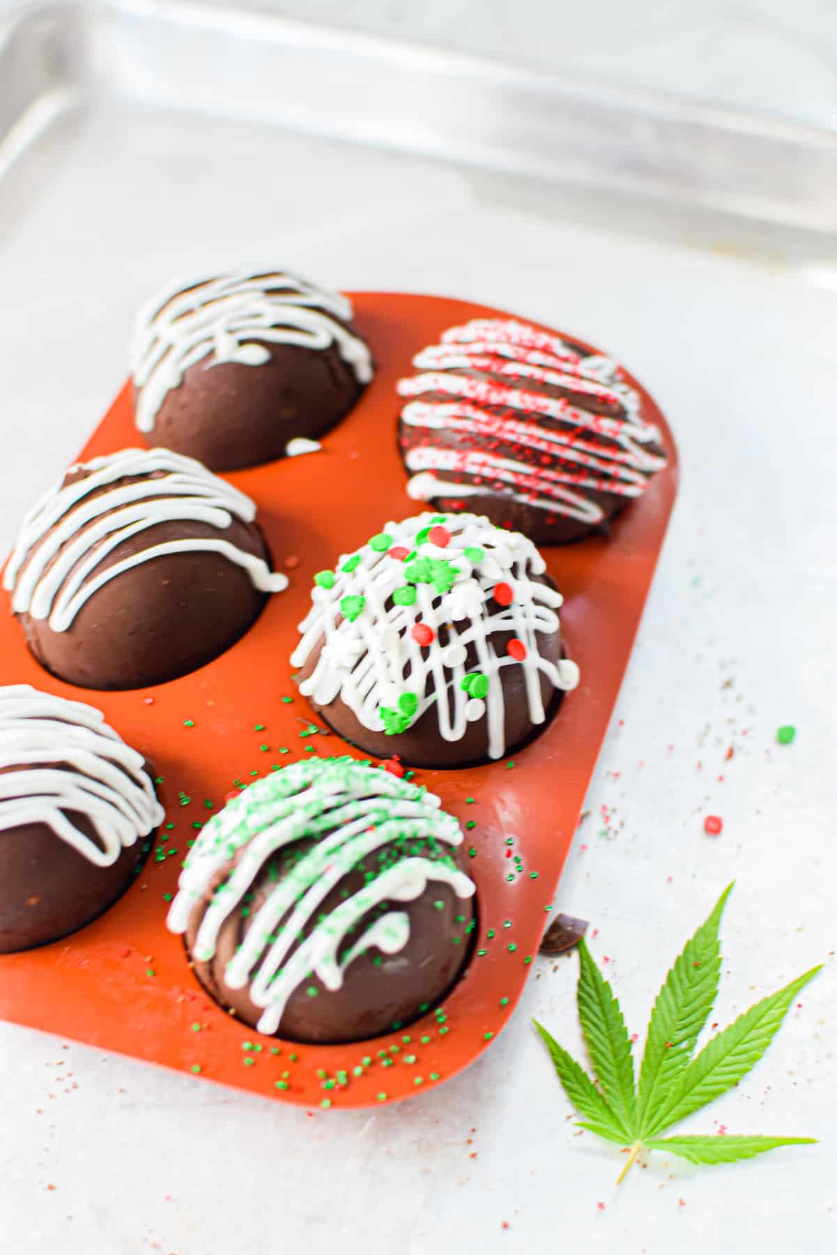 A parchment lined baking sheet with a mold full of completed hot chocolate bombs garnished with white chocolate and sprinkles, with a cannabis leaf on the side