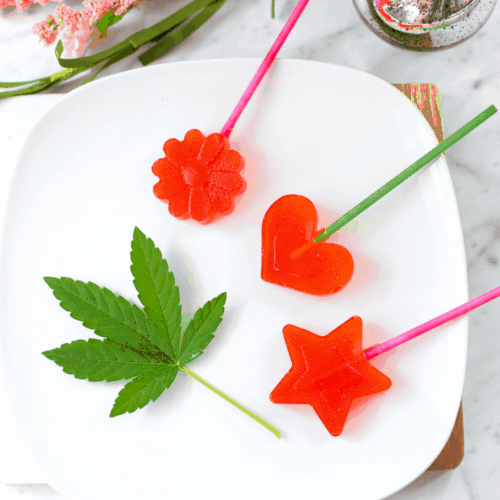 A white plate with 3 red cannabis lollipops laying on it with a cannabis leaf on the left side for garnish