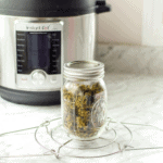 A picture of a white counter top with an instant pot in the background focusing on a mason jar of cannabis flowers on a trivet