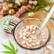 A white countertop with a cutting board topped with a clear mug full of cannabis hot cocoa with marshmallows, garnished with a cannabis leaf