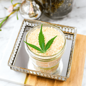 A white counter top with a cutting board with a mirrored tray topped with a mason jar full of cannabis salt, garnished with a cannabis leaf