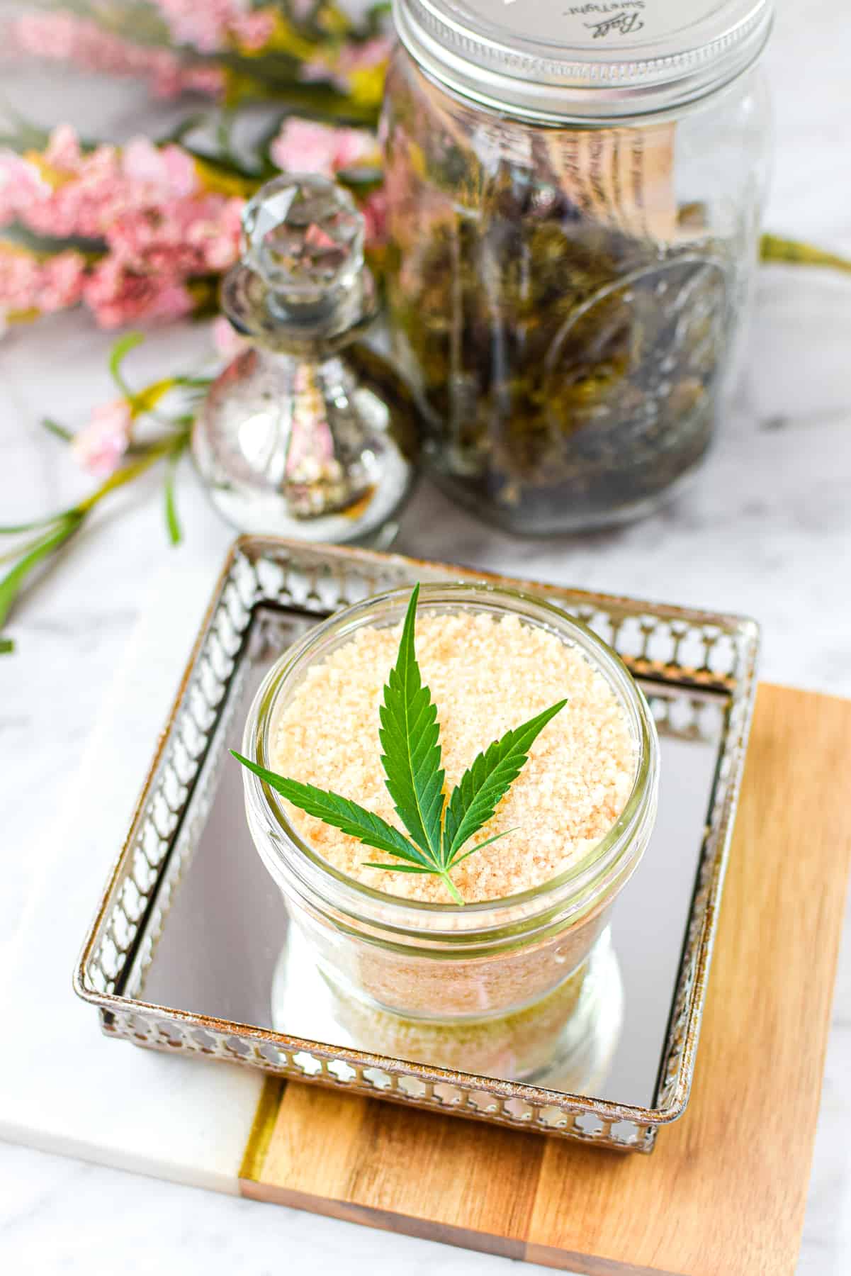 A white counter top with a cutting board with a mirrored tray topped with a mason jar full of cannabis salt, garnished with a cannabis leaf