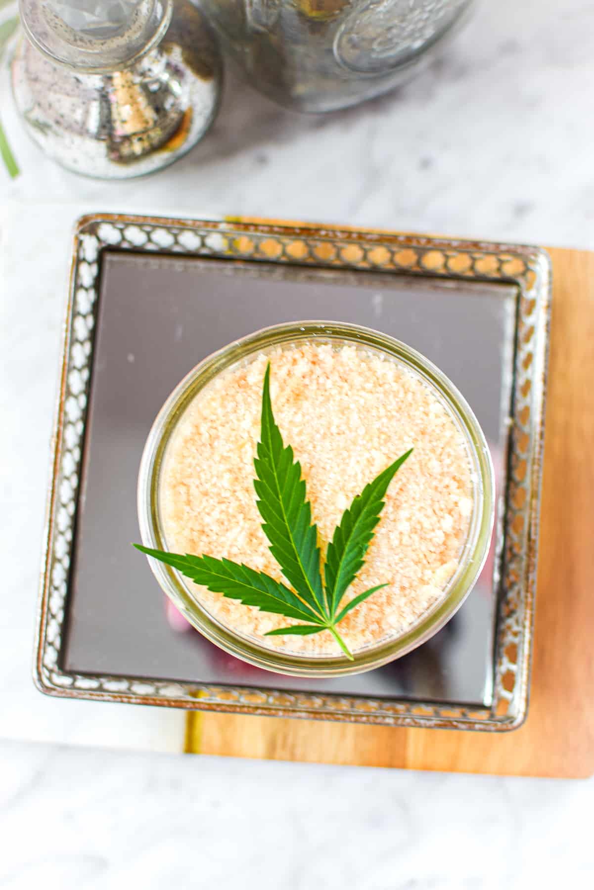 A white counter top with a cutting board with a mirrored tray topped with a mason jar full of cannabis salt, garnished with a cannabis leaf