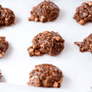 A white countertop with a cutting board topped with a white plate topped with three no-bake rocky road cannabis cookies