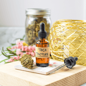 A white countertop with a brown tincture bottle labeled THCA tincture with a cannabis flower bud next to it with a hand gripping the top of the tincture dropper
