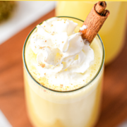 A white countertop with a cutting board topped with two glasses of cannabis eggnog topped with whipped cream