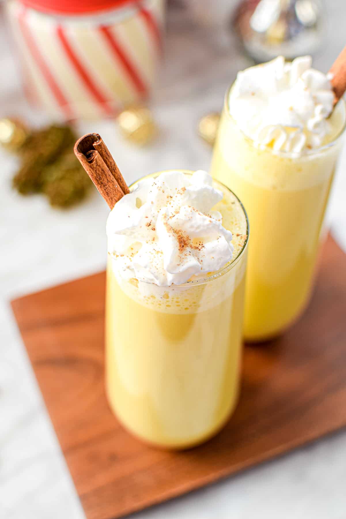 A white countertop with a cutting board topped with two glasses of cannabis eggnog topped with whipped cream