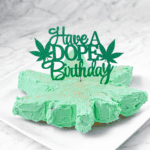 A white countertop and a white plate with a green frosted cannabis cake with a topper that says have a dope birthday