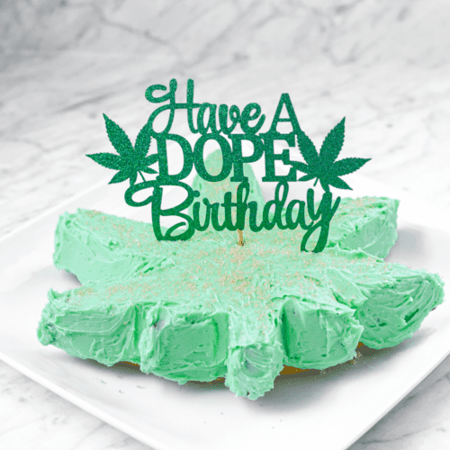 A white countertop and a white plate with a green frosted cannabis cake with a topper that says have a dope birthday
