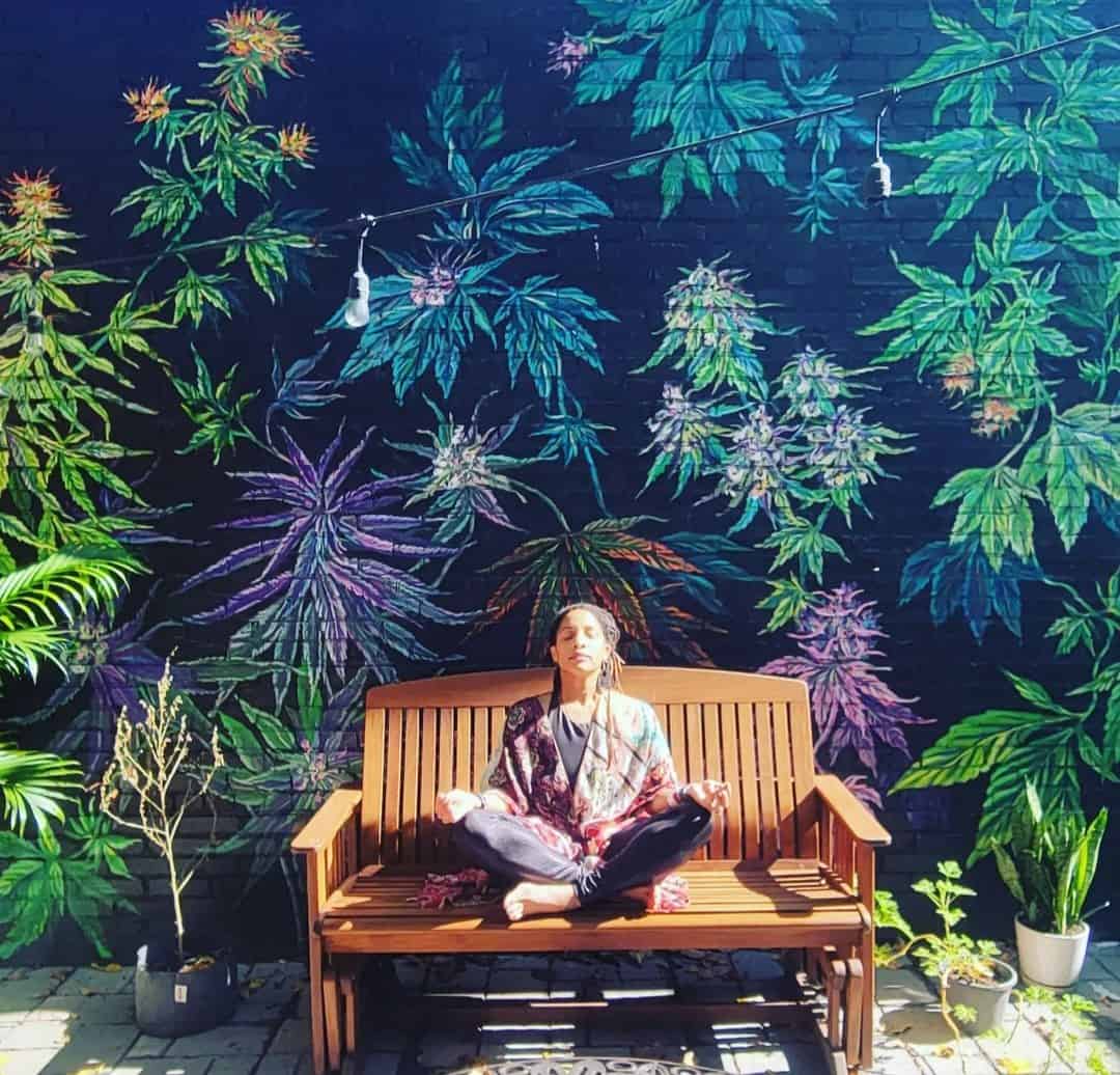 A picture of Sharyn Lustre sitting on a wooden bench in front of a cannabis mural.