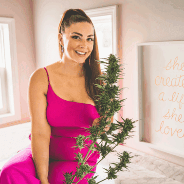 A woman in a pink dress holding a cannabis plant, smiling, promoting DIY cannabis drying room tips and home cultivation advice.