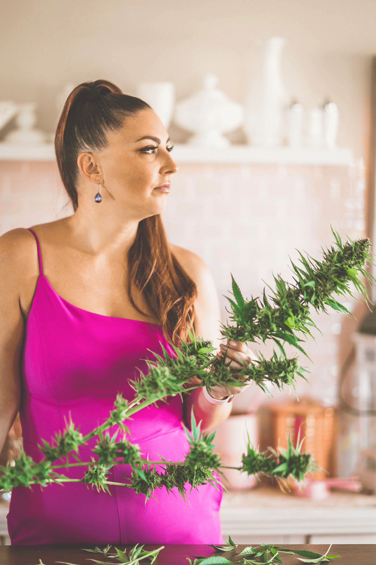 A woman in a pink dress holding a cannabis plant, smiling, promoting DIY cannabis drying room tips and home cultivation advice.